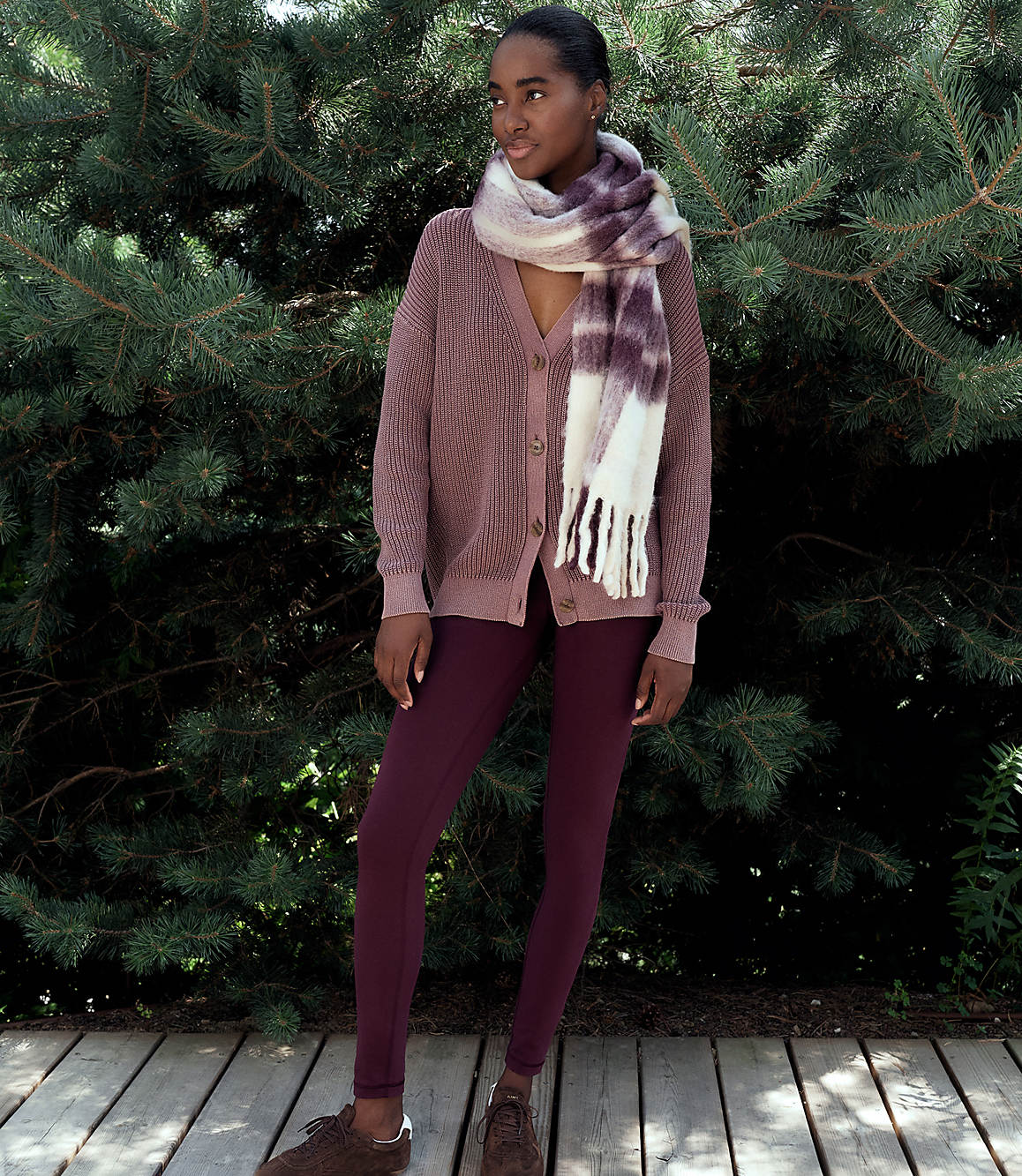 A woman wearing a mauve cardigan, burgundy leggings, and a patterned scarf stands on a wooden deck against a backdrop of dense evergreen trees.