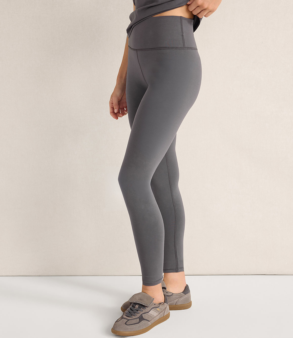 A person wearing grey leggings and sneakers stands against a plain, light-colored wall.