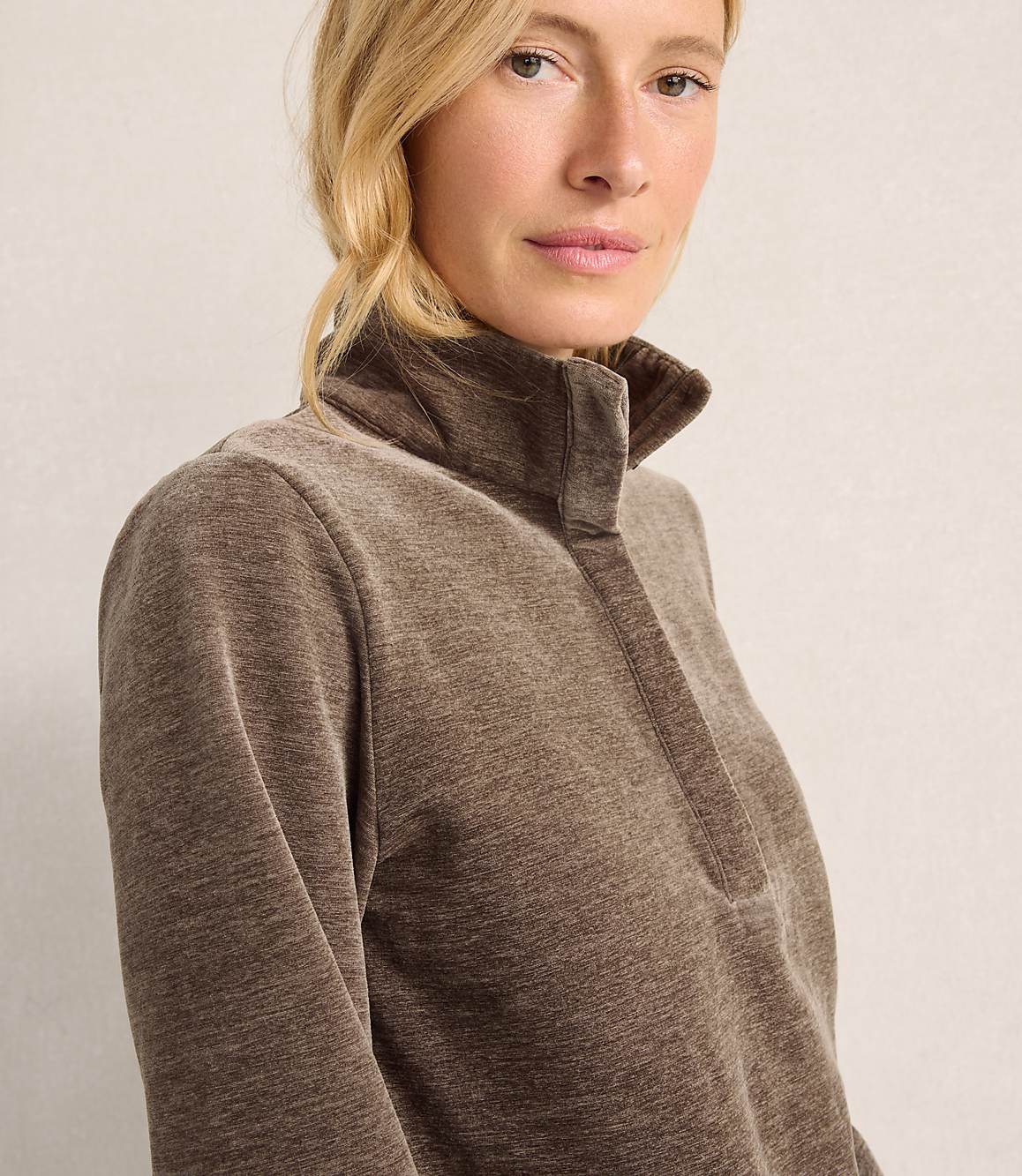 A blonde woman wearing a brown quarter-zip pullover stands against a plain, light-colored background.