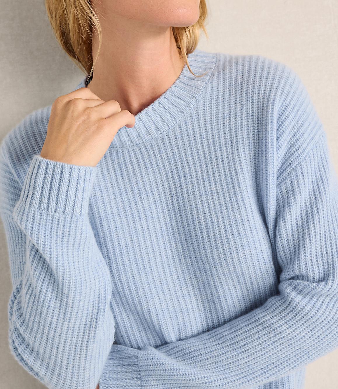 A person wearing a light blue ribbed sweater stands against a plain, neutral-toned wall.