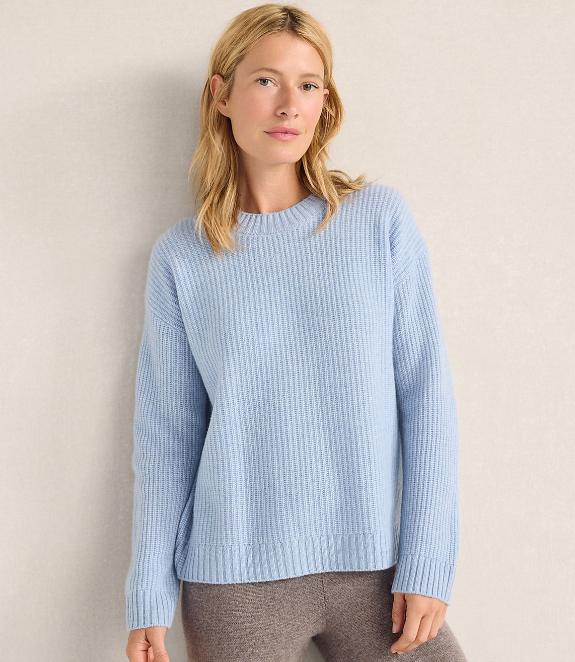 A blonde woman wearing a light blue ribbed sweater and brown pants stands against a plain, neutral-toned wall.