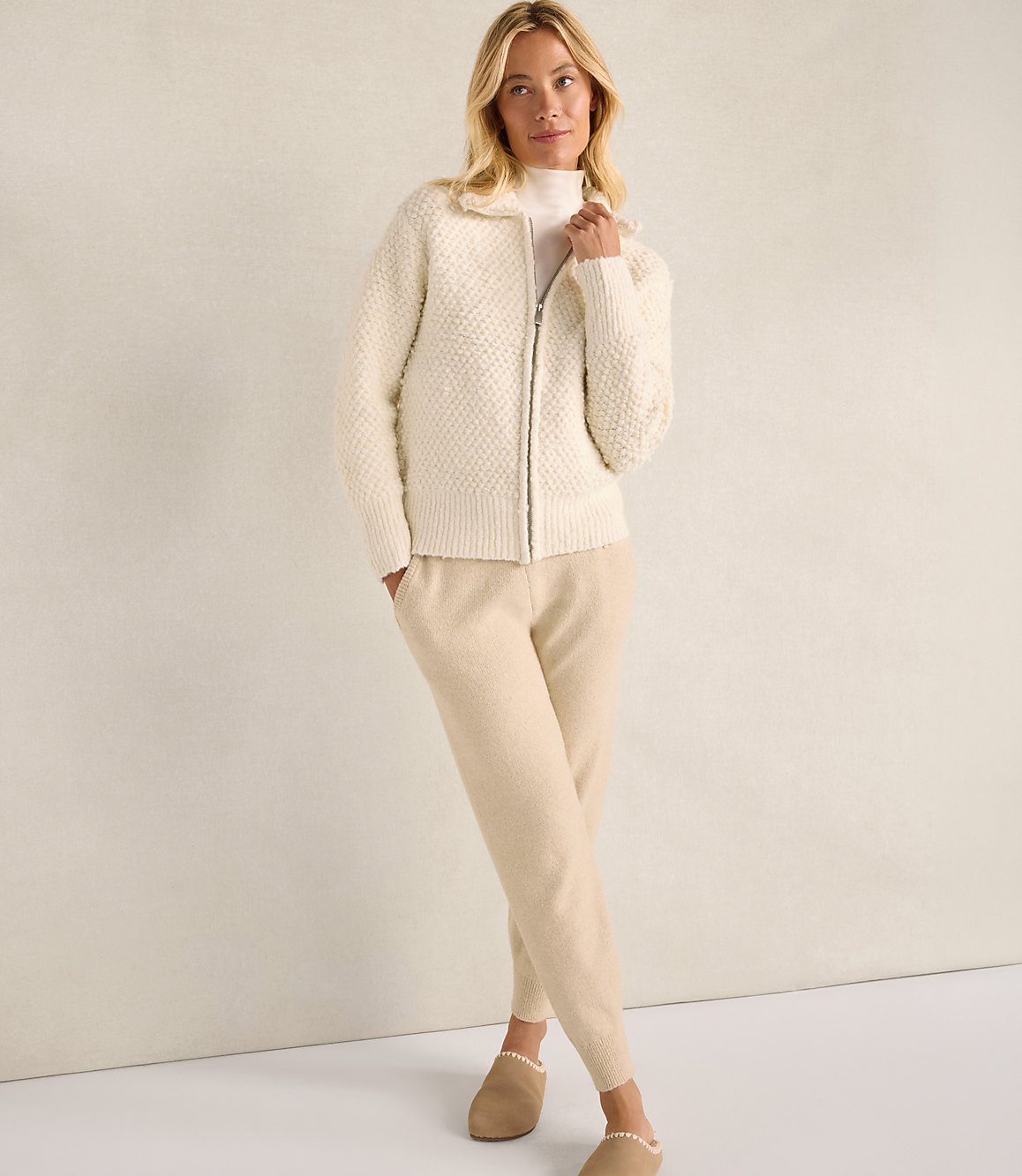 A woman wearing a cream-colored textured zip-up jacket and matching pants stands against a plain, neutral-toned wall.