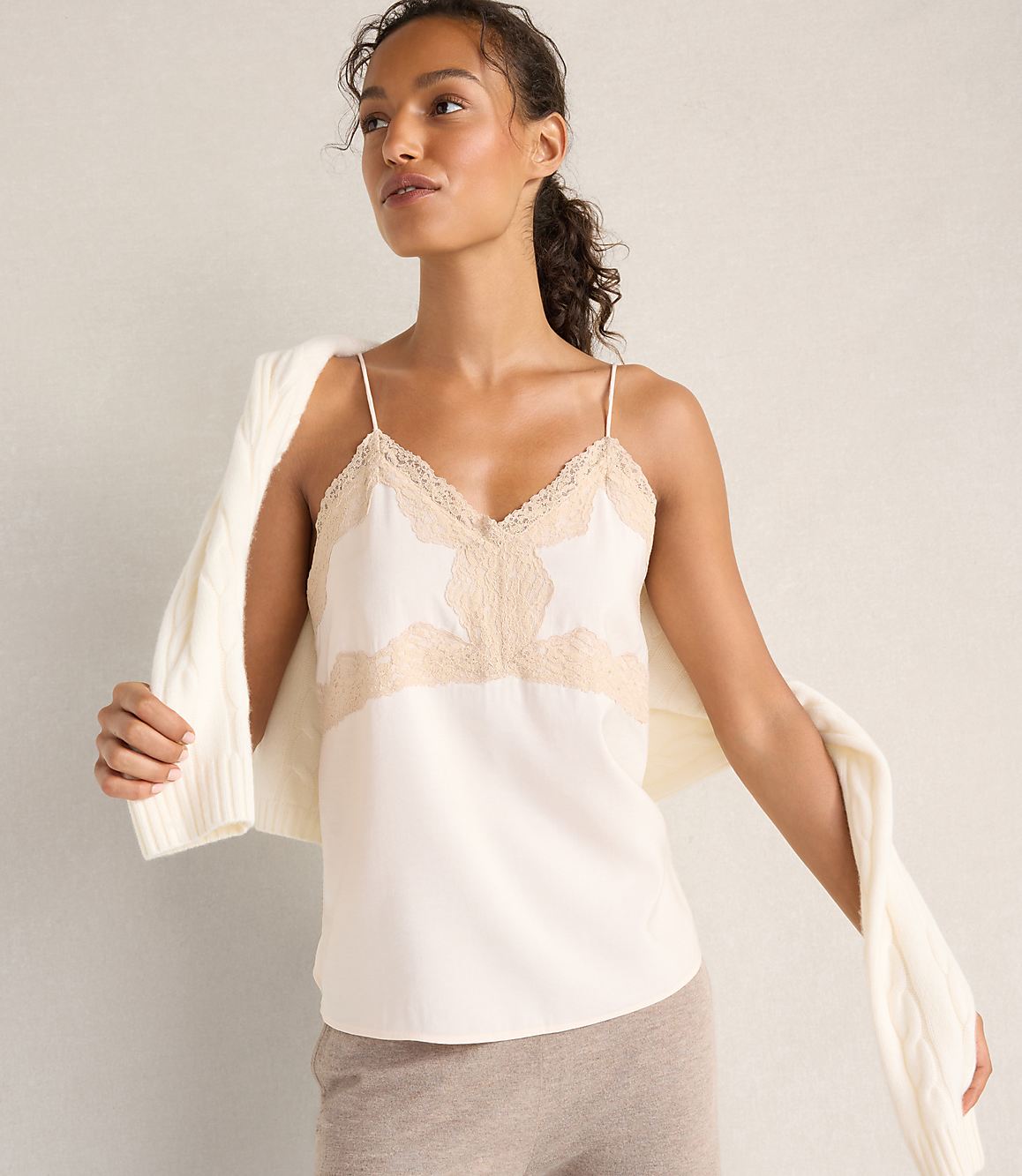 A woman wearing a white lace-trimmed camisole and a white cardigan draped over her shoulders stands against a plain, neutral-toned wall.