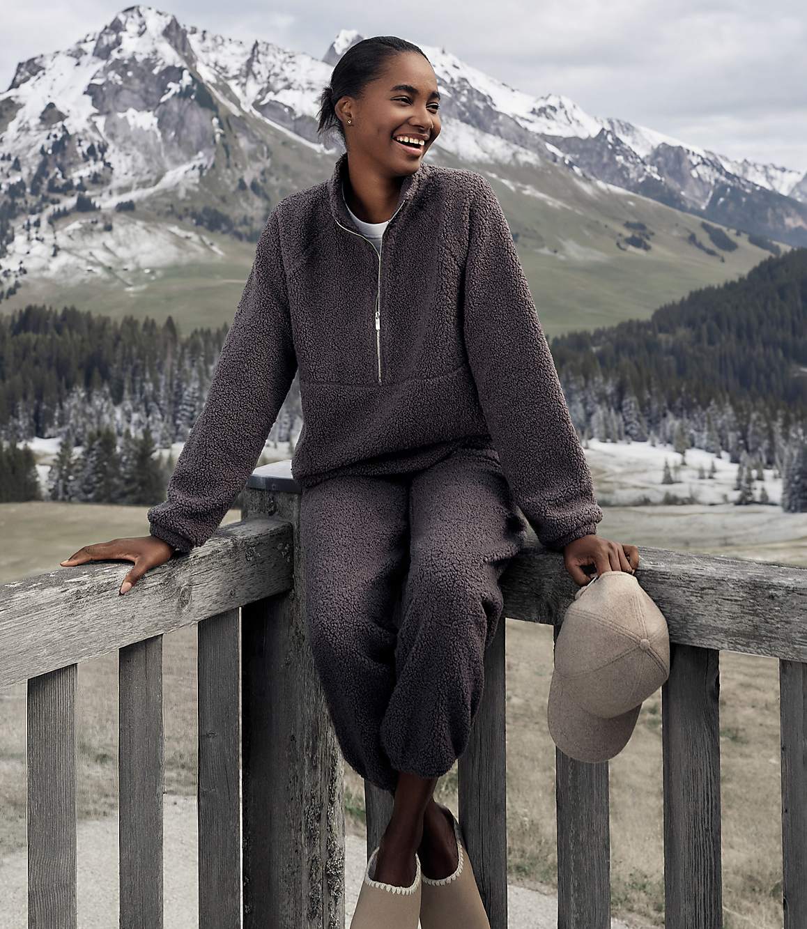 A smiling woman in a charcoal fleece set sits on a wooden railing against a backdrop of snow-capped mountains and evergreen forests.