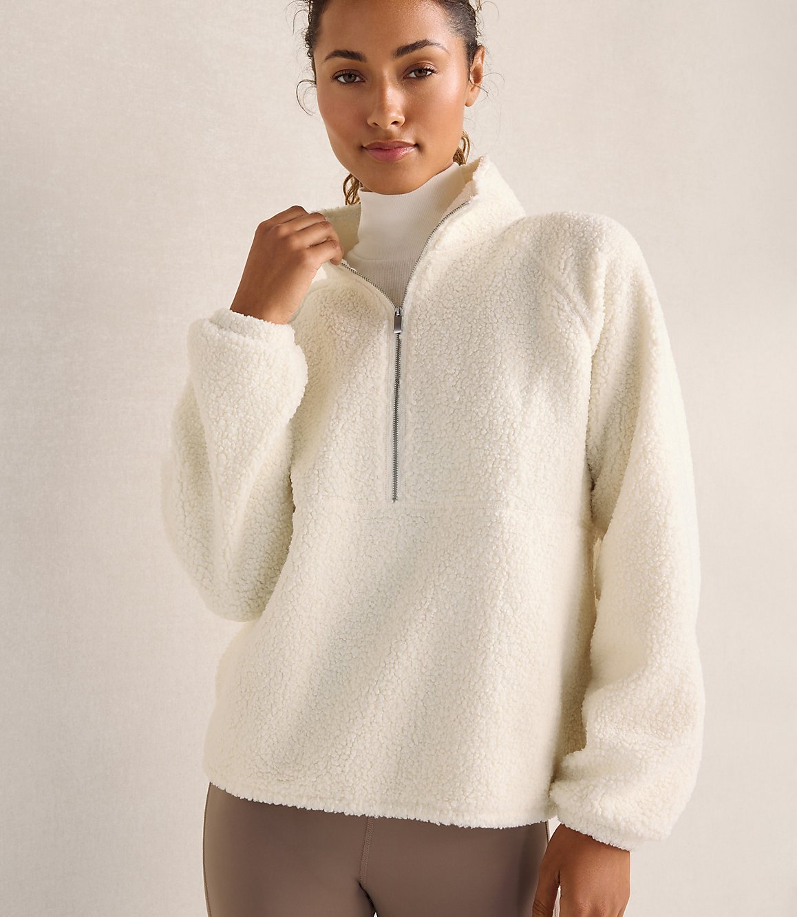 A woman wearing a white fleece pullover and a white turtleneck stands against a plain, neutral-toned background.