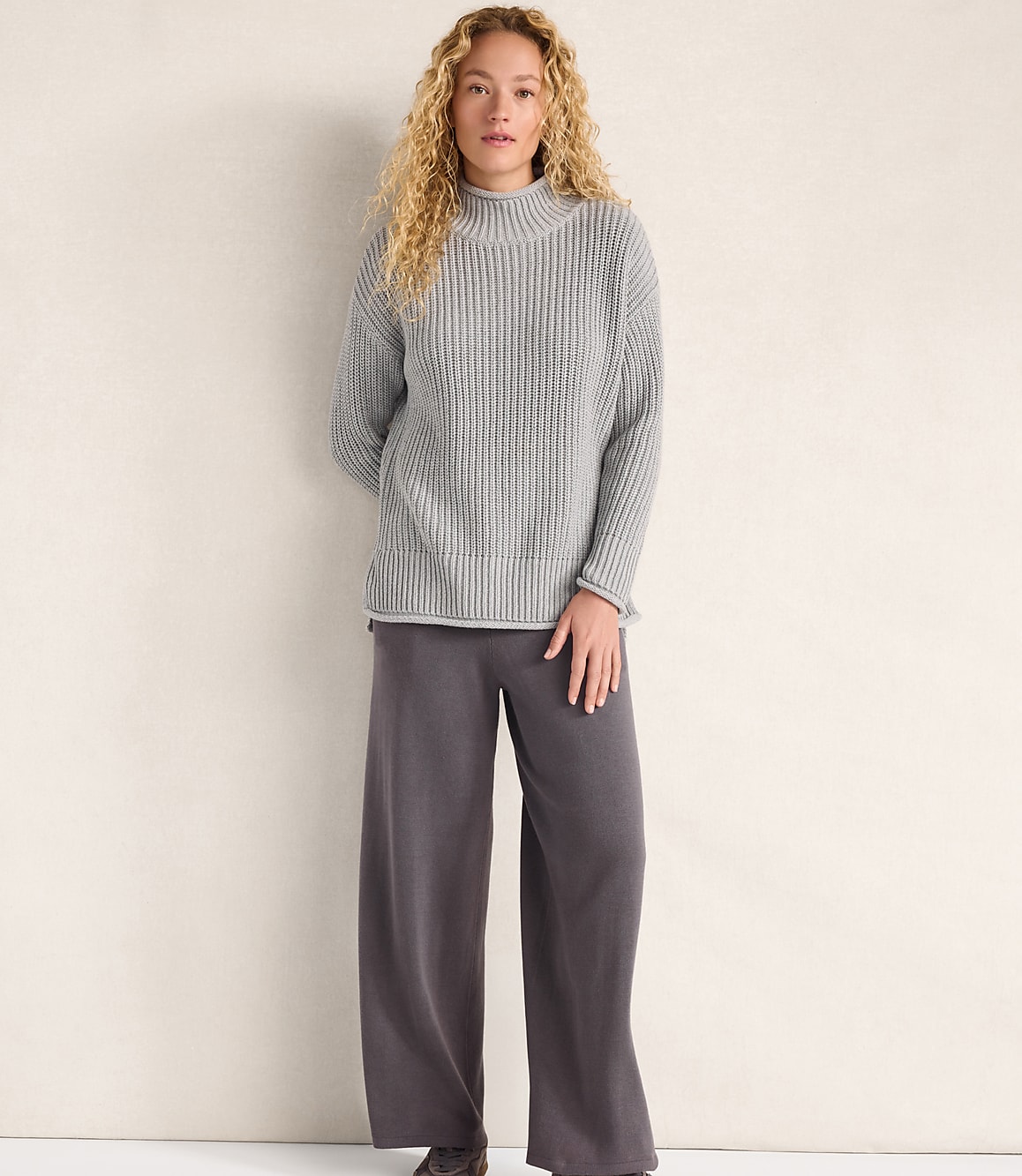 A woman with curly blonde hair wearing a grey ribbed sweater and wide-leg charcoal trousers stands against a plain, light-colored wall.