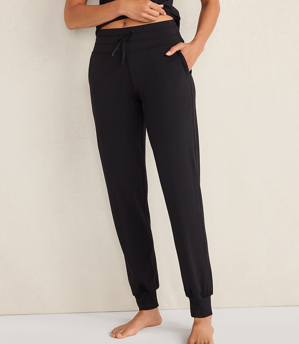 A person wearing black jogger pants stands against a plain, light-colored wall.