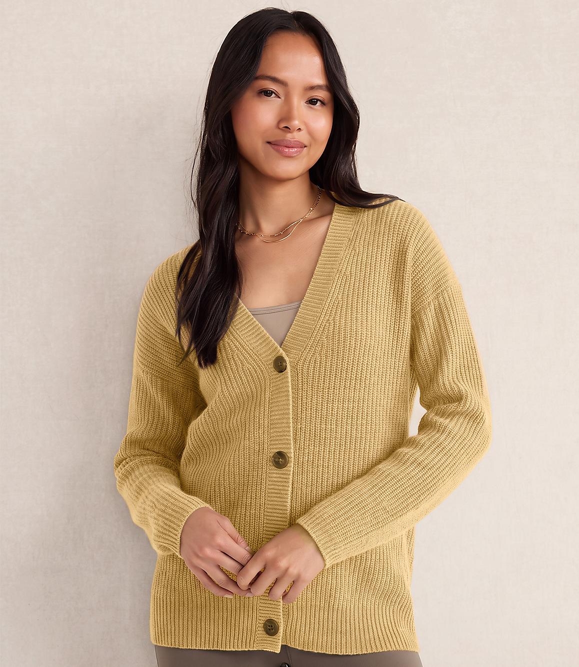 A woman wearing a yellow ribbed cardigan and gold necklaces stands against a plain, light-colored wall.