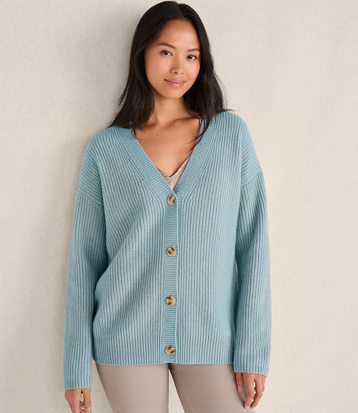 A woman wearing a light blue ribbed cardigan and beige pants stands against a plain, neutral-toned wall.
