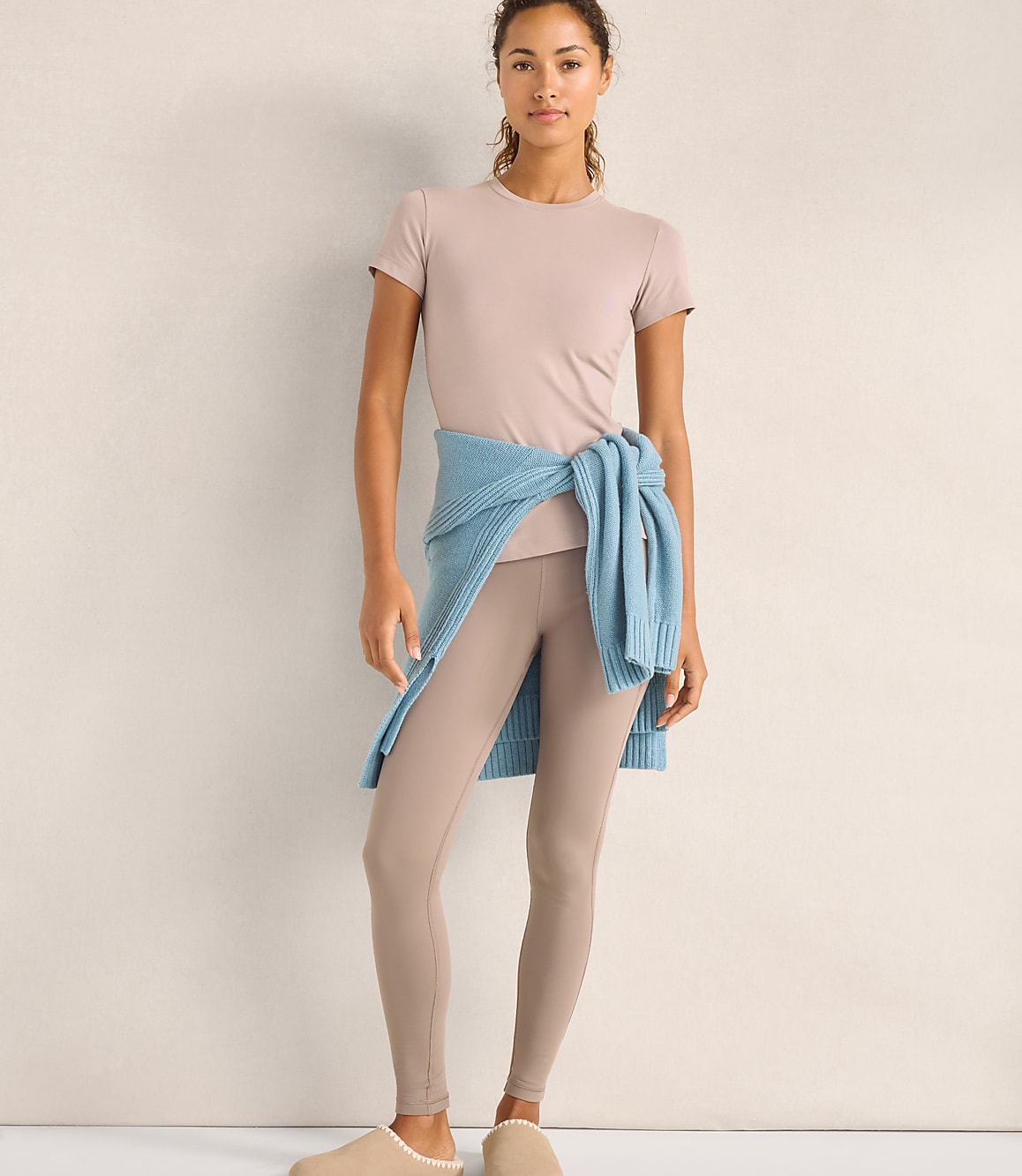 A woman wearing a beige t-shirt and leggings with a blue sweater tied around her waist stands against a plain, neutral-colored wall.