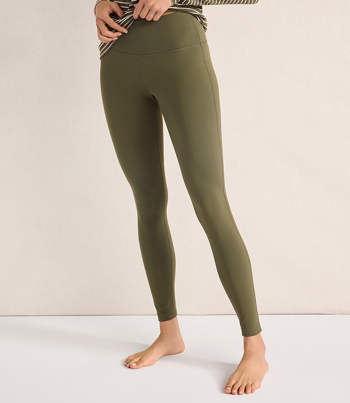 A person wearing olive green leggings and a striped shirt stands against a plain, light-colored background.
