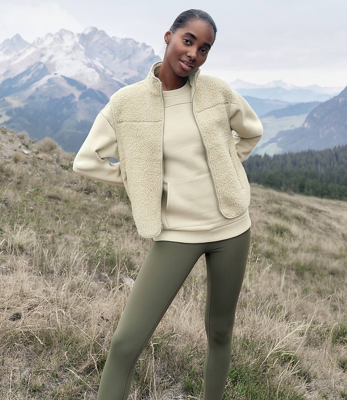 A woman wearing a cream-colored fleece vest and olive green leggings stands in a grassy field with snow-capped mountains in the background.