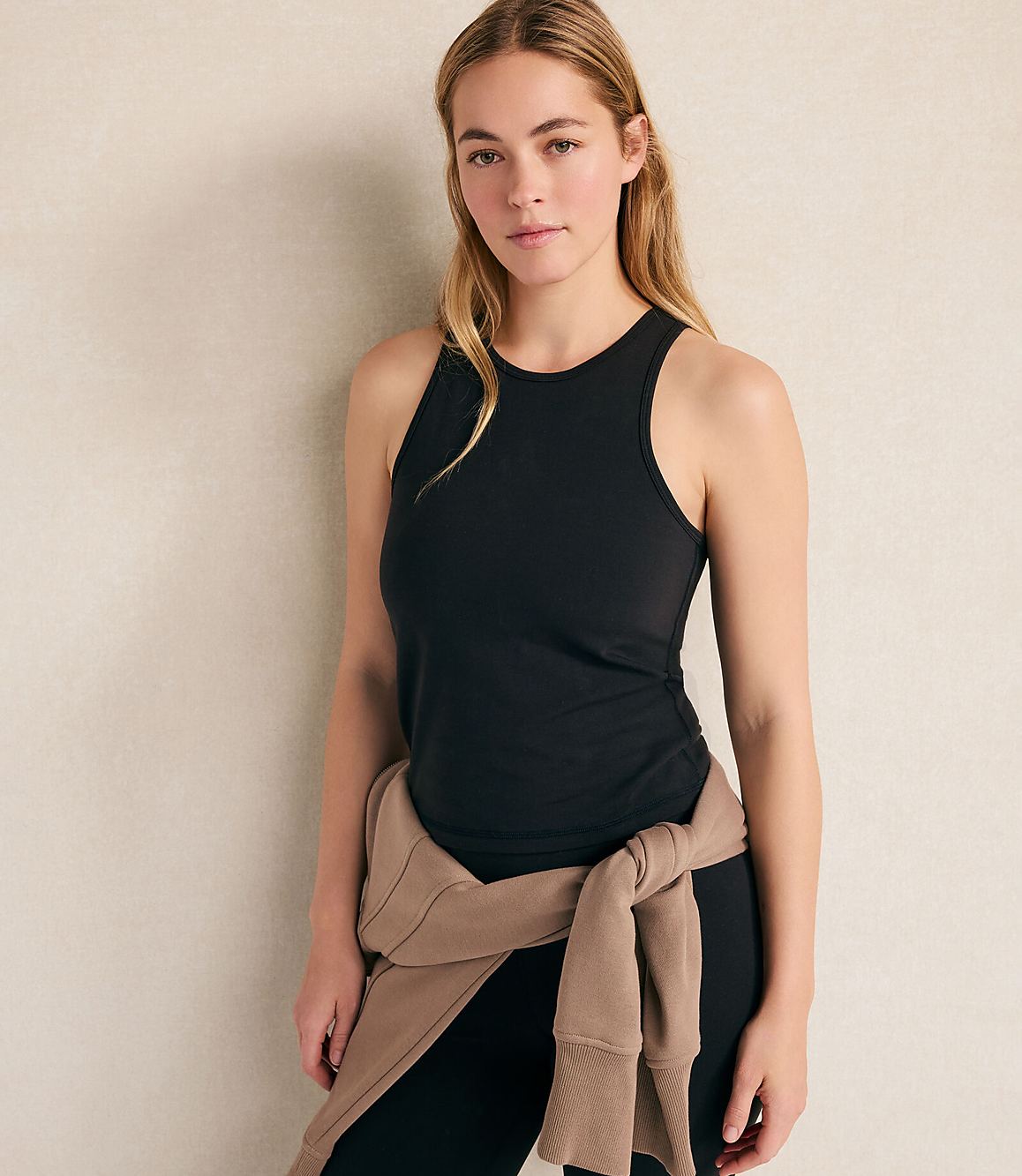 A woman wearing a black tank top and leggings with a tan sweater tied around her waist stands against a plain, light-colored wall.