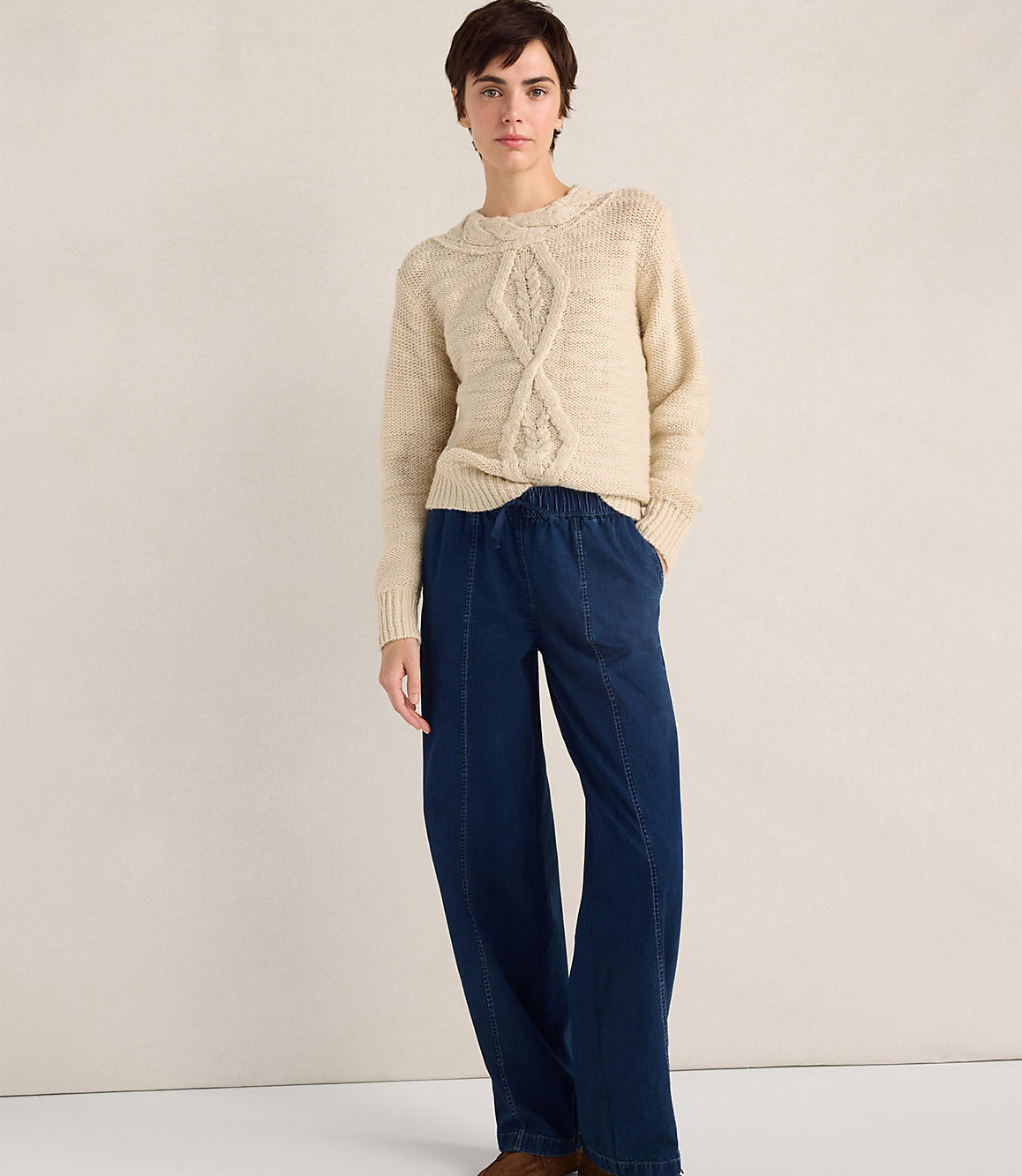 A person wearing a cream cable-knit sweater and wide-leg navy blue trousers stands against a plain, neutral-colored wall.