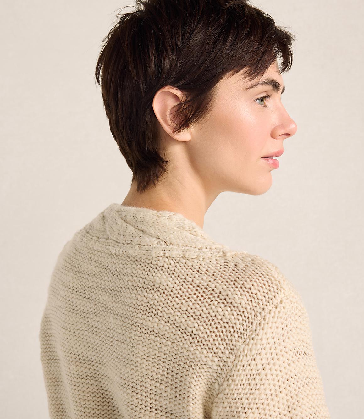 A woman with short dark hair wearing a beige knit sweater stands against a plain, light-colored background.