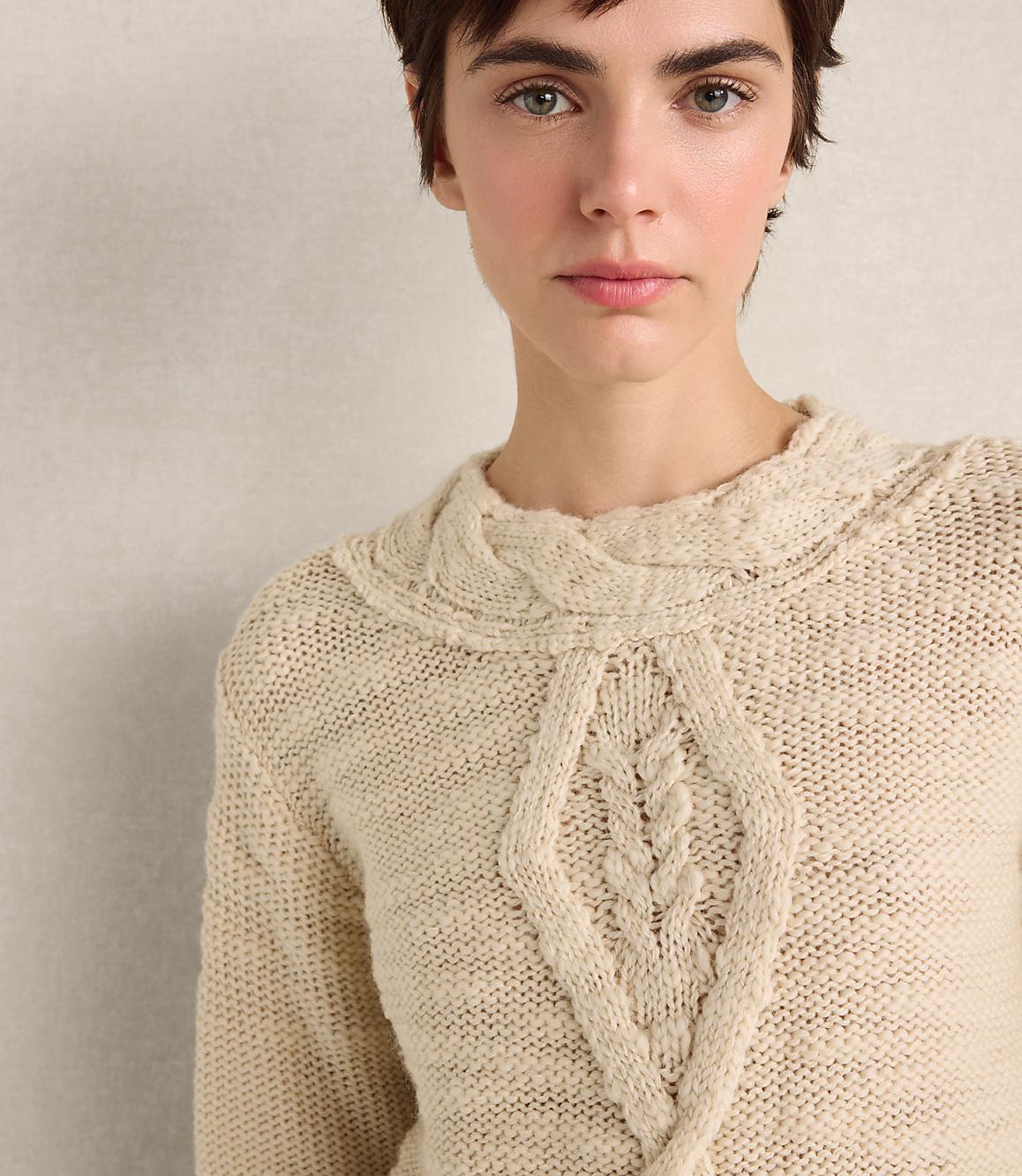 A woman with short dark hair wearing a cream-colored cable-knit sweater stands against a plain, neutral-toned wall.
