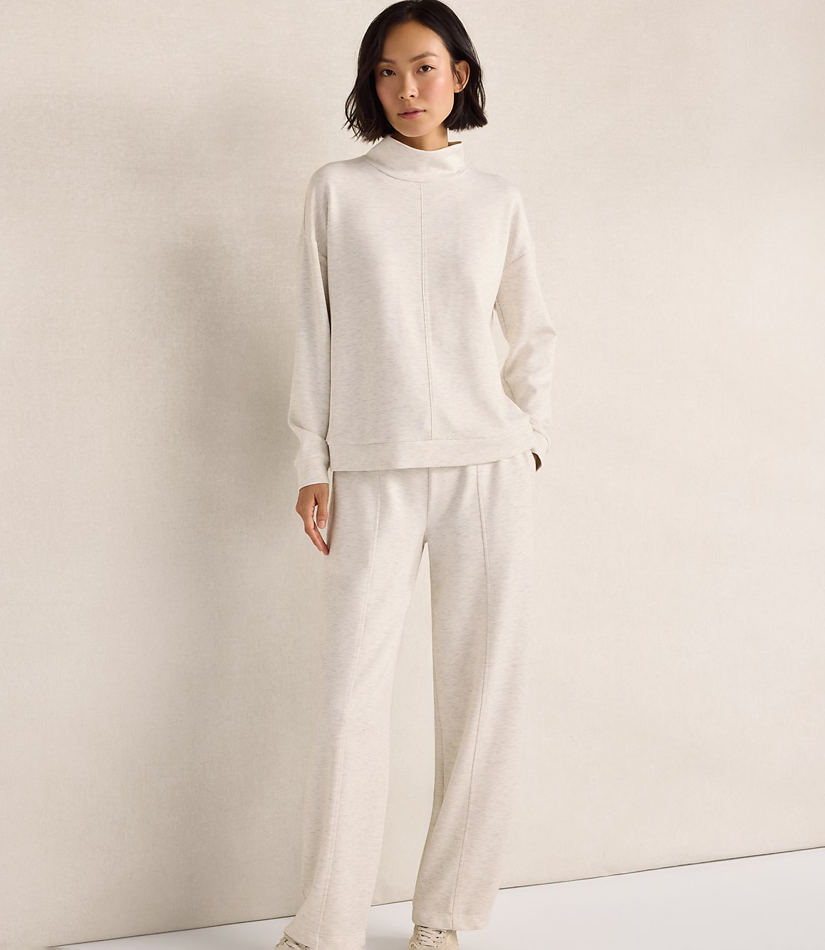 A woman wearing a cream-colored matching sweater and pants set stands against a plain, light-colored wall.