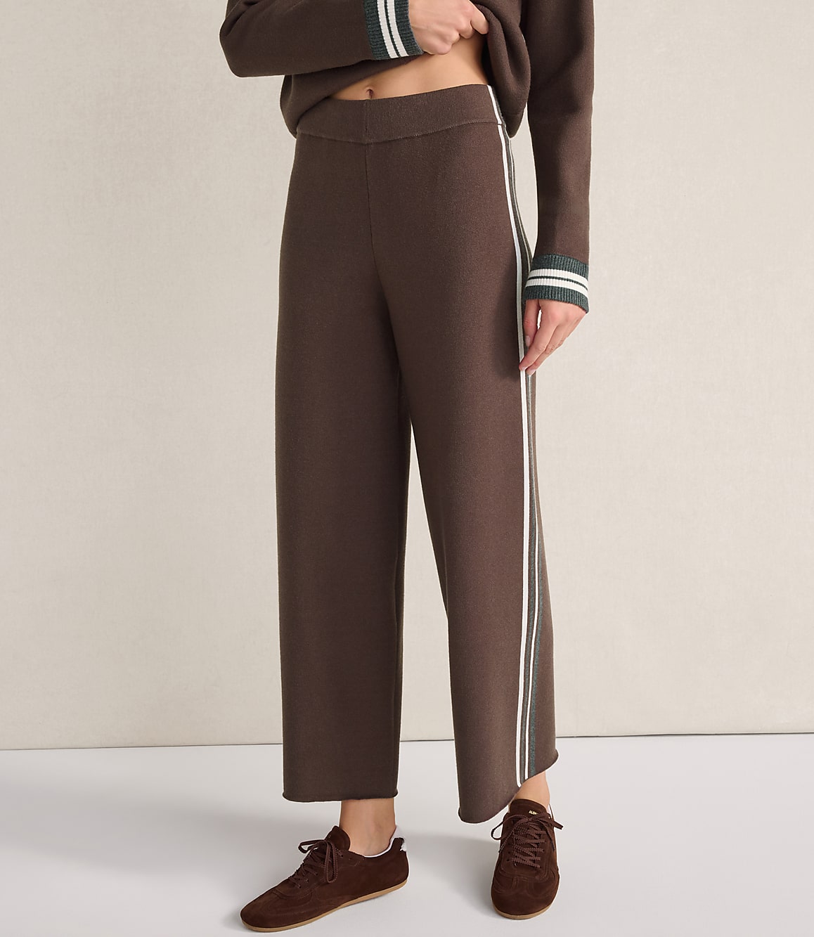 A person wears brown wide-leg trousers with white side stripes and matching brown shoes against a plain, light-colored background.