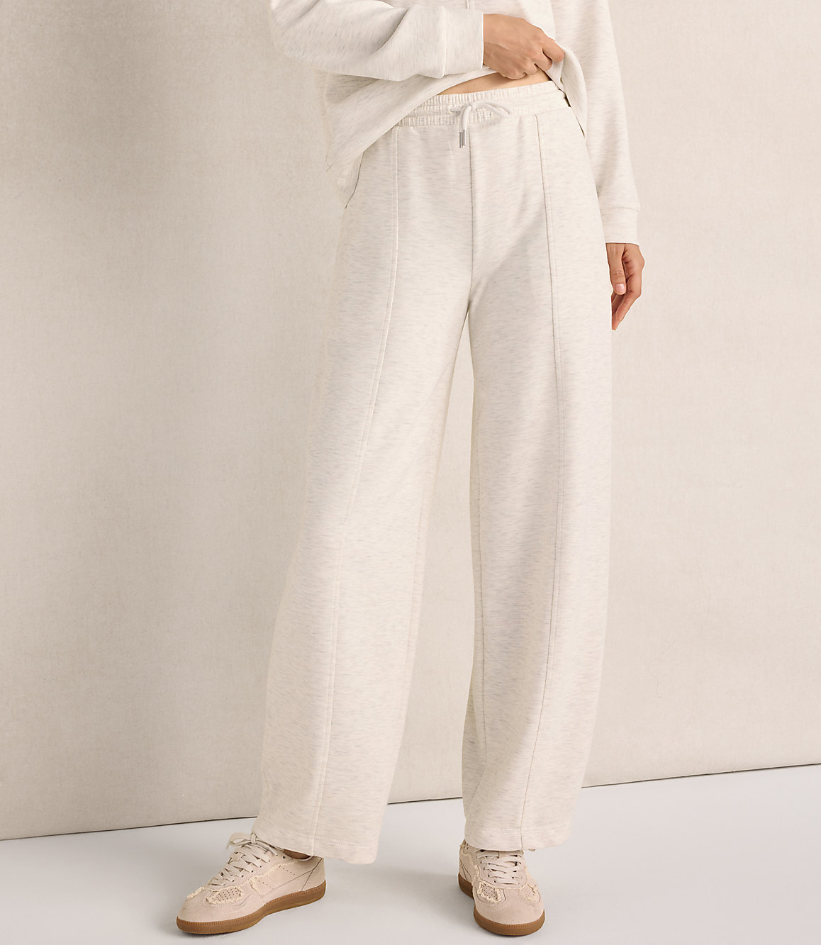 A person wearing a cream-colored sweatshirt and wide-leg lounge pants stands against a neutral, light-beige wall.
