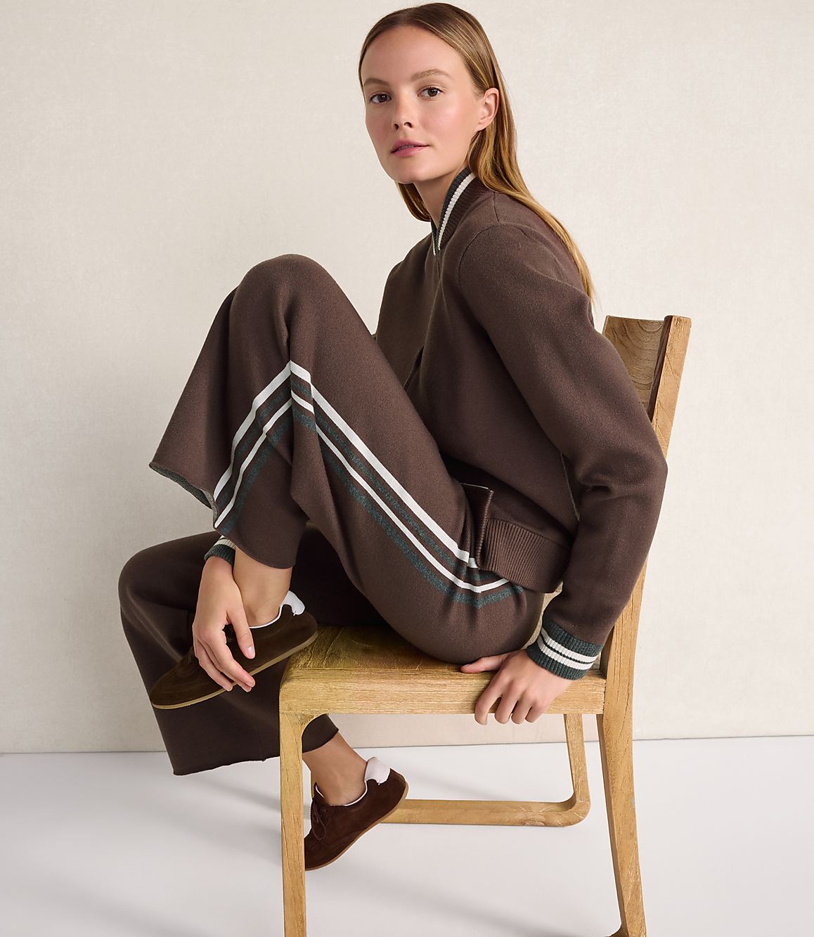 A woman wearing a brown tracksuit and loafers sits on a wooden chair against a plain, light-colored wall.
