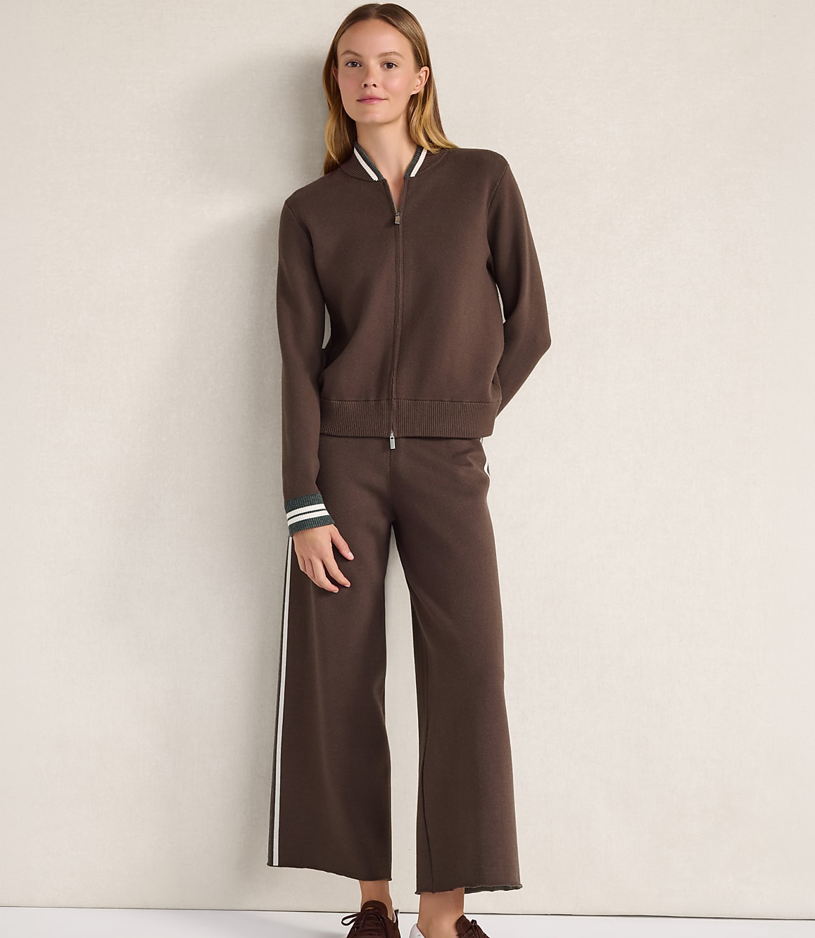 A woman wearing a brown zip-up jacket and matching wide-leg trousers stands against a plain, light-colored wall.