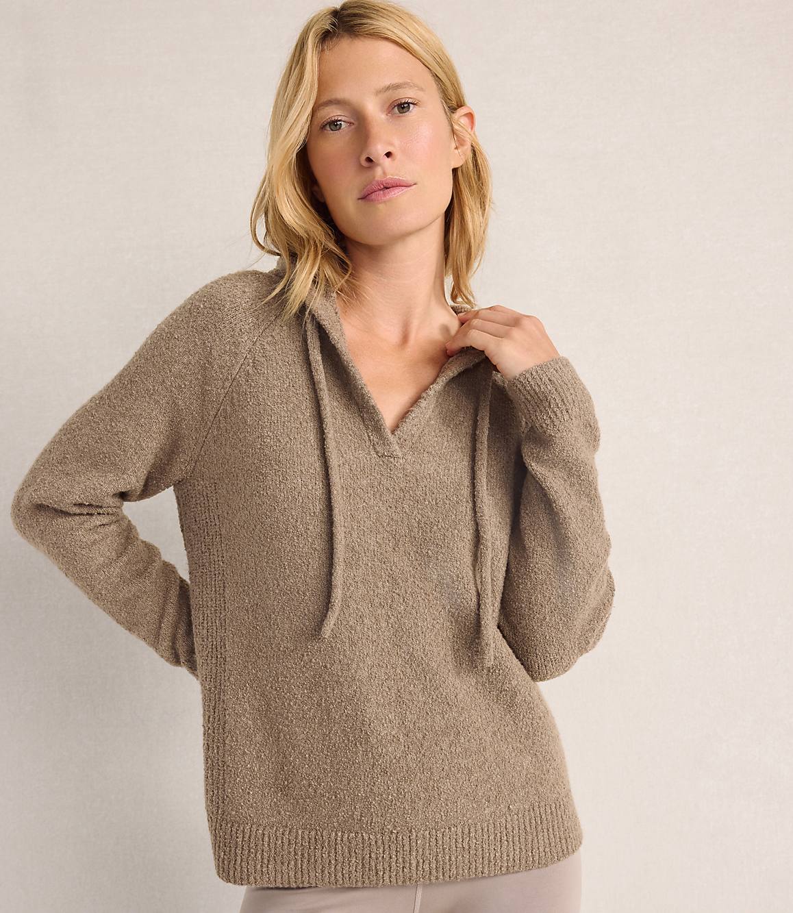 A blonde woman wearing a taupe knit sweater stands against a plain, light-colored background.