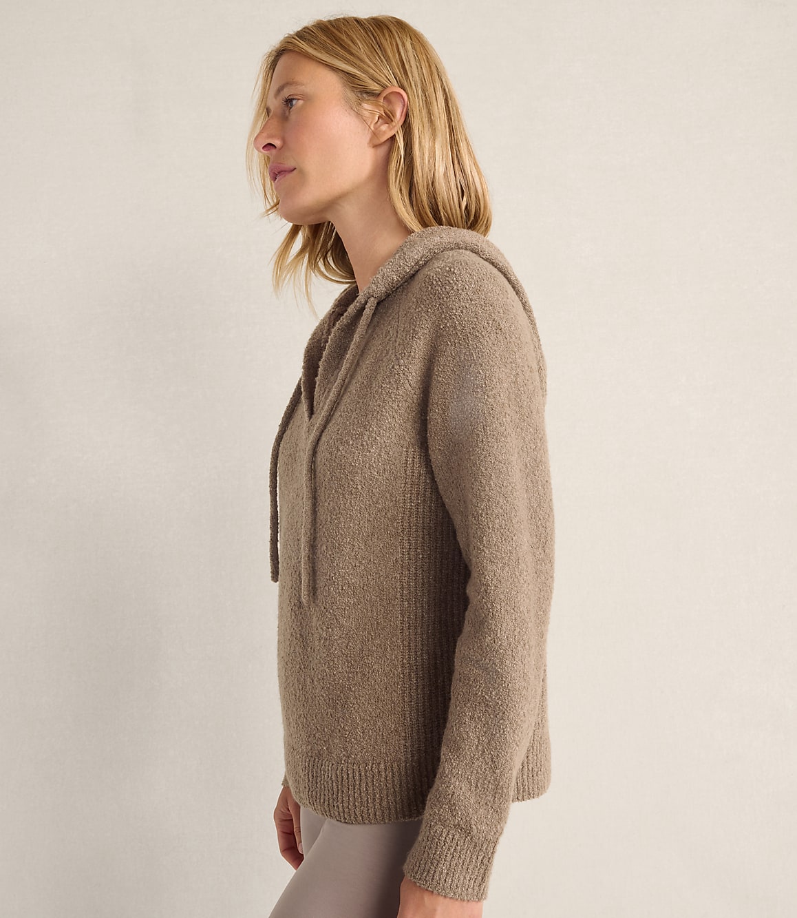A blonde woman wearing a taupe hooded sweater stands in profile against a plain, light-colored wall.