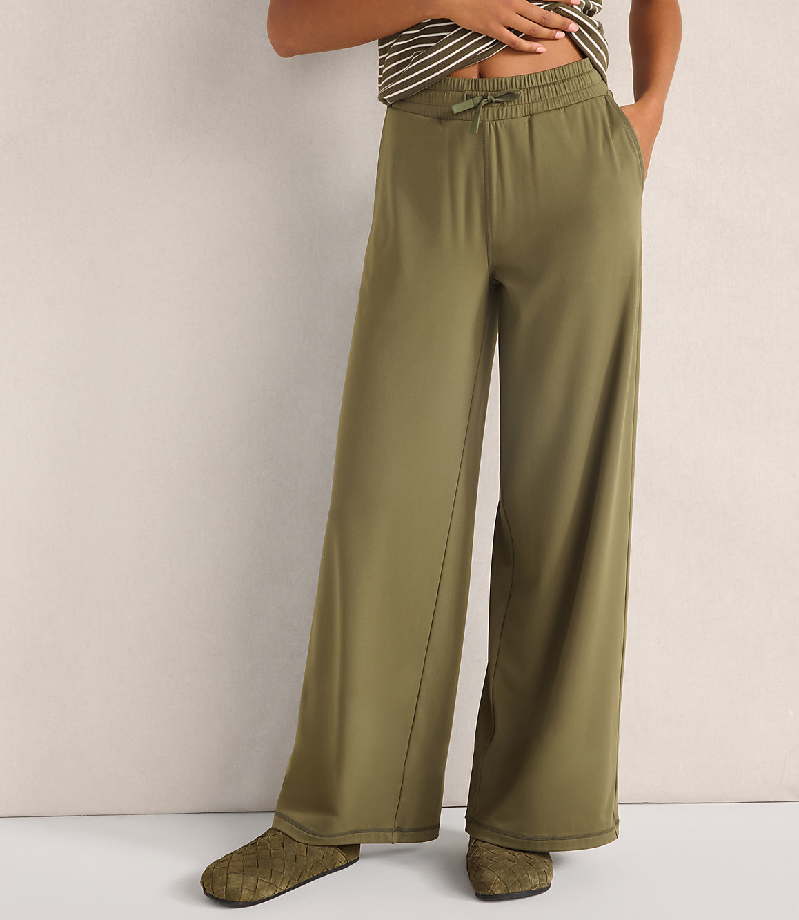 A person wears olive green wide-leg trousers and woven flats against a plain, neutral-toned wall.
