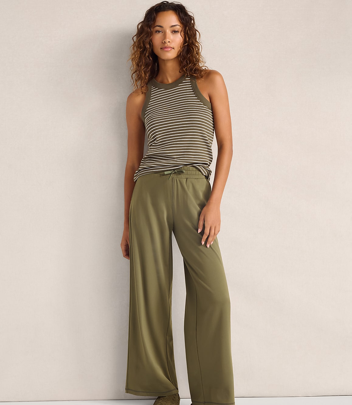 A woman wearing a striped tank top and olive green wide-leg pants stands against a plain, neutral-colored wall.