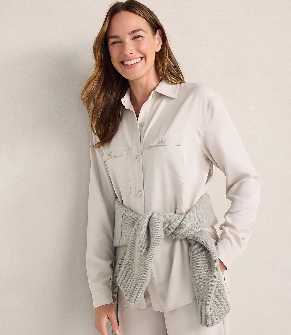 A smiling woman wearing a light-colored button-down shirt with a grey sweater tied around her waist stands against a plain, neutral-toned wall.
