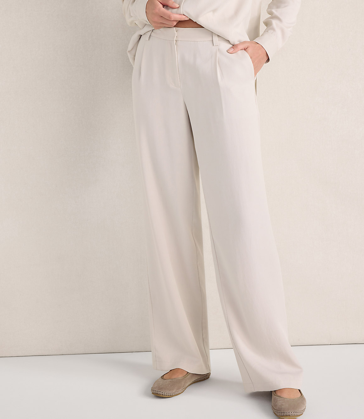A person wears wide-leg cream trousers and beige flats against a plain, light-colored wall.