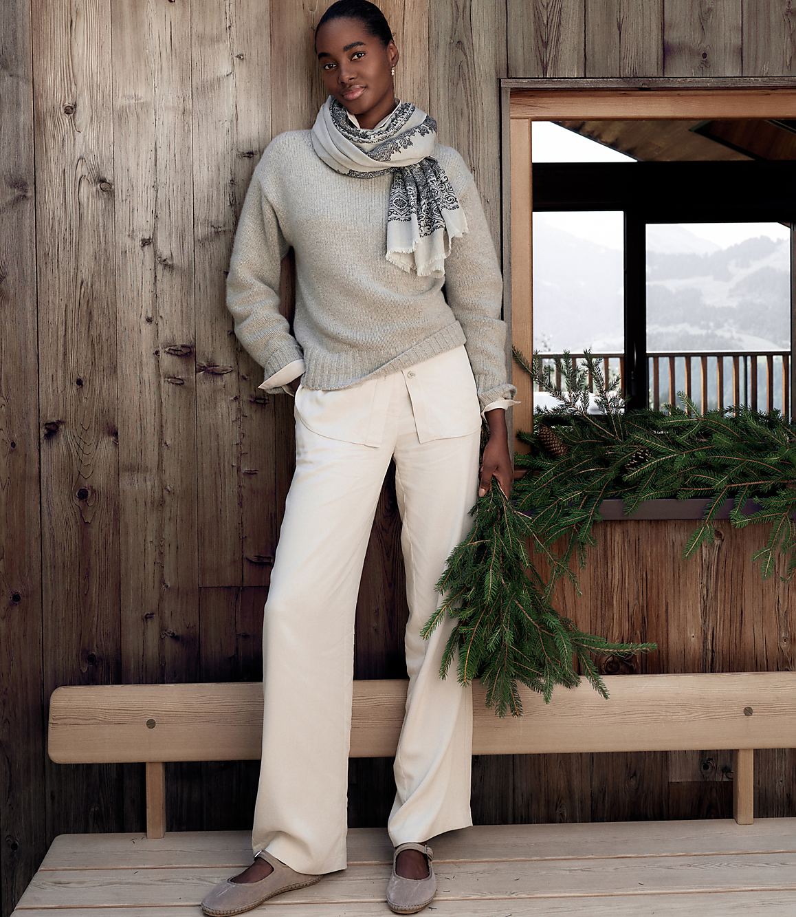 A woman wearing a grey sweater, scarf, and white trousers stands before a rustic wooden wall and window overlooking a snowy landscape.