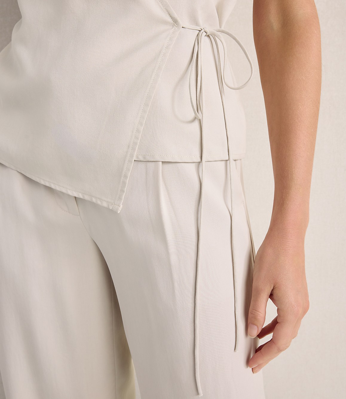 A person wears a cream-colored wrap top and matching wide-leg trousers against a neutral, textured background.