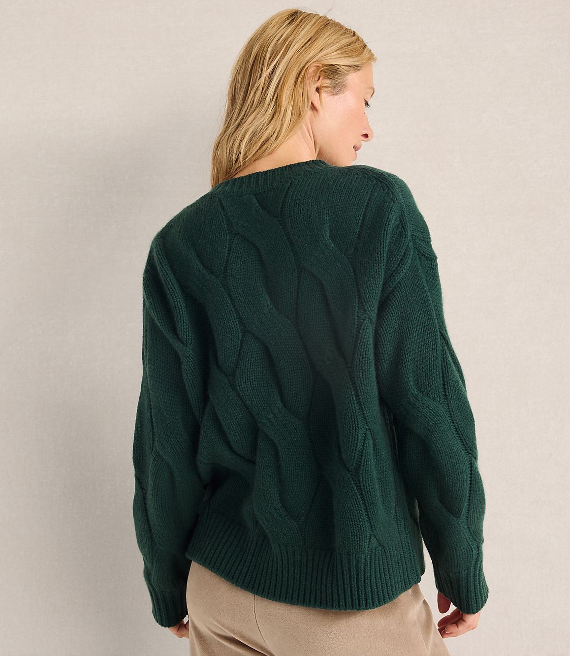 A woman with blonde hair wearing a dark green cable-knit sweater and tan pants stands against a plain, light-colored wall.