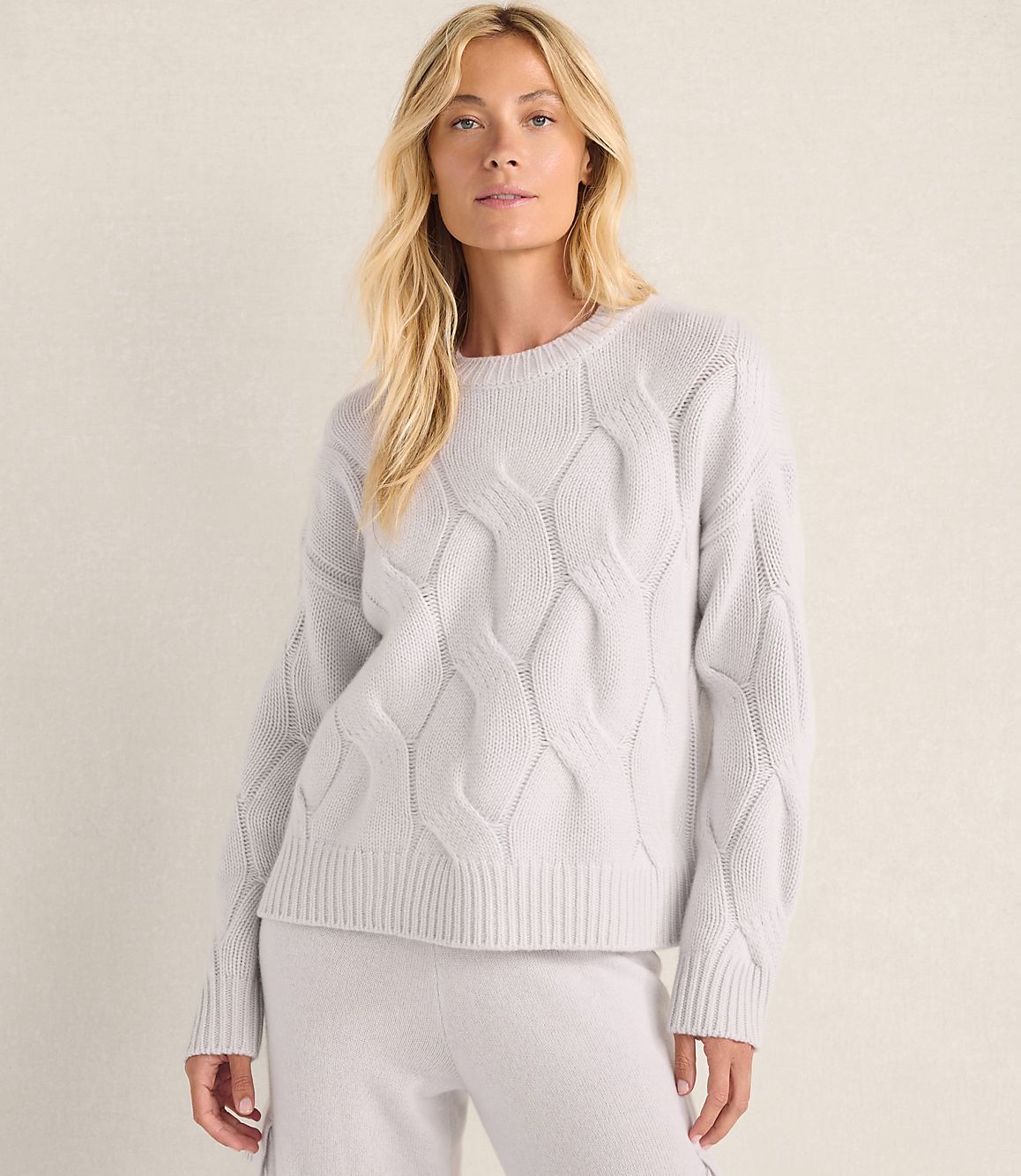 A blonde woman wearing a light purple cable-knit sweater and matching pants stands against a plain, neutral-colored wall.