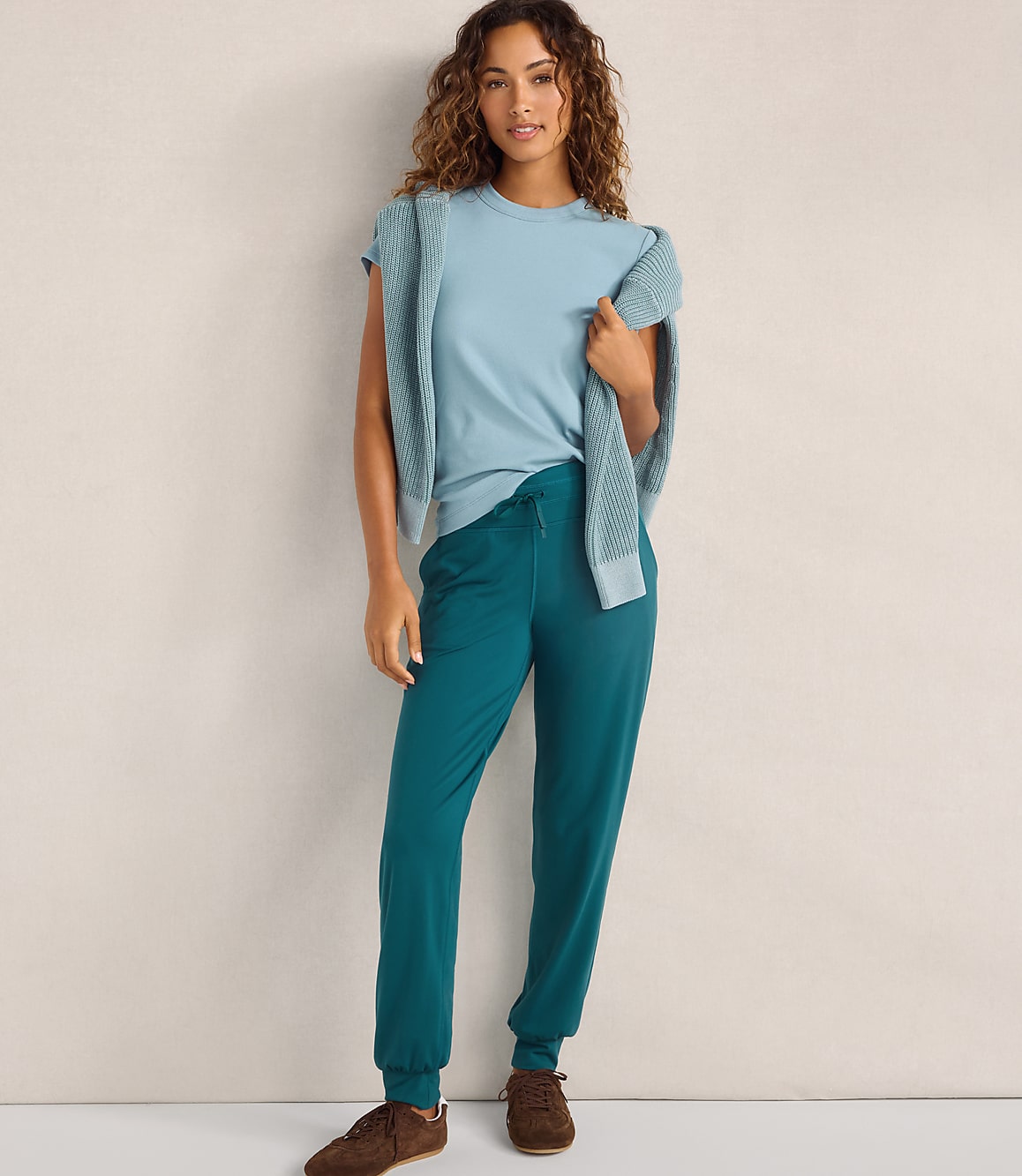 A woman wearing a light blue t-shirt, a matching cardigan, and teal joggers stands against a plain, neutral-colored wall.