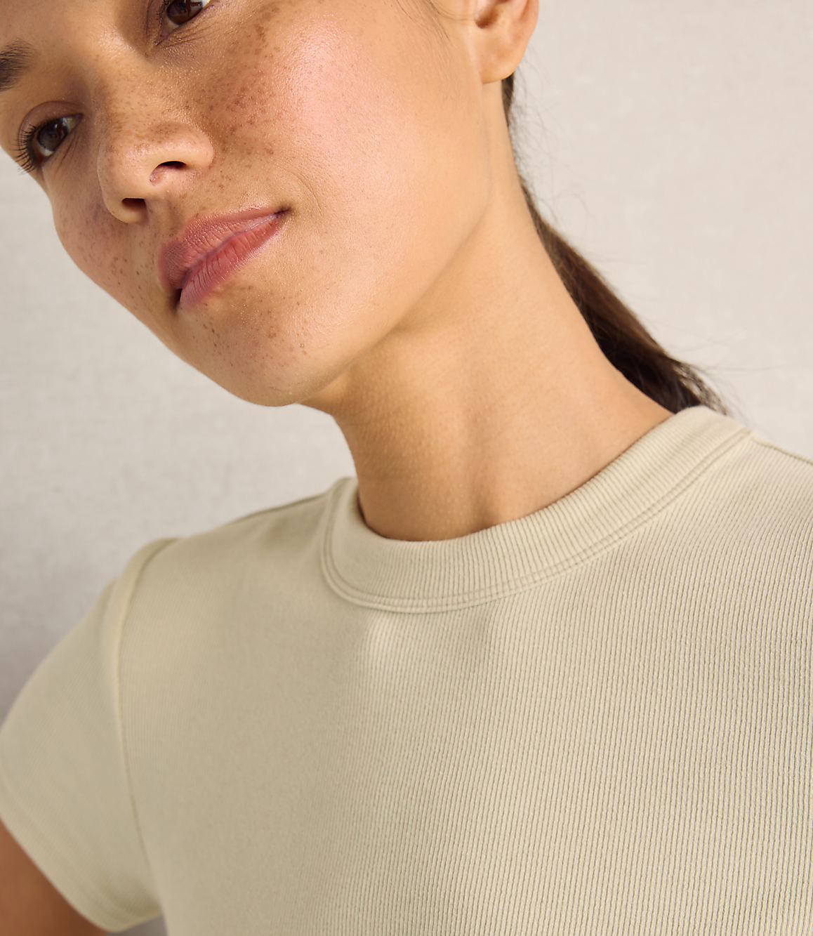 A close-up of a woman with freckles wearing a beige ribbed t-shirt against a plain, light-colored wall.