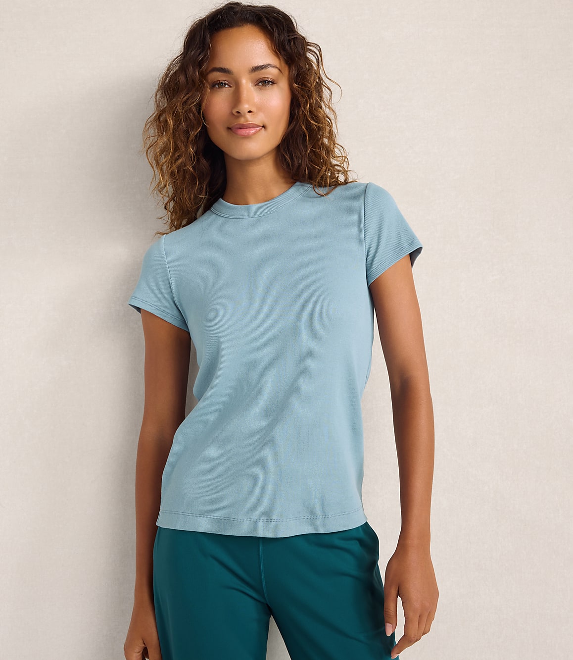 A woman wearing a light blue t-shirt and dark teal pants stands against a plain, neutral-toned wall.