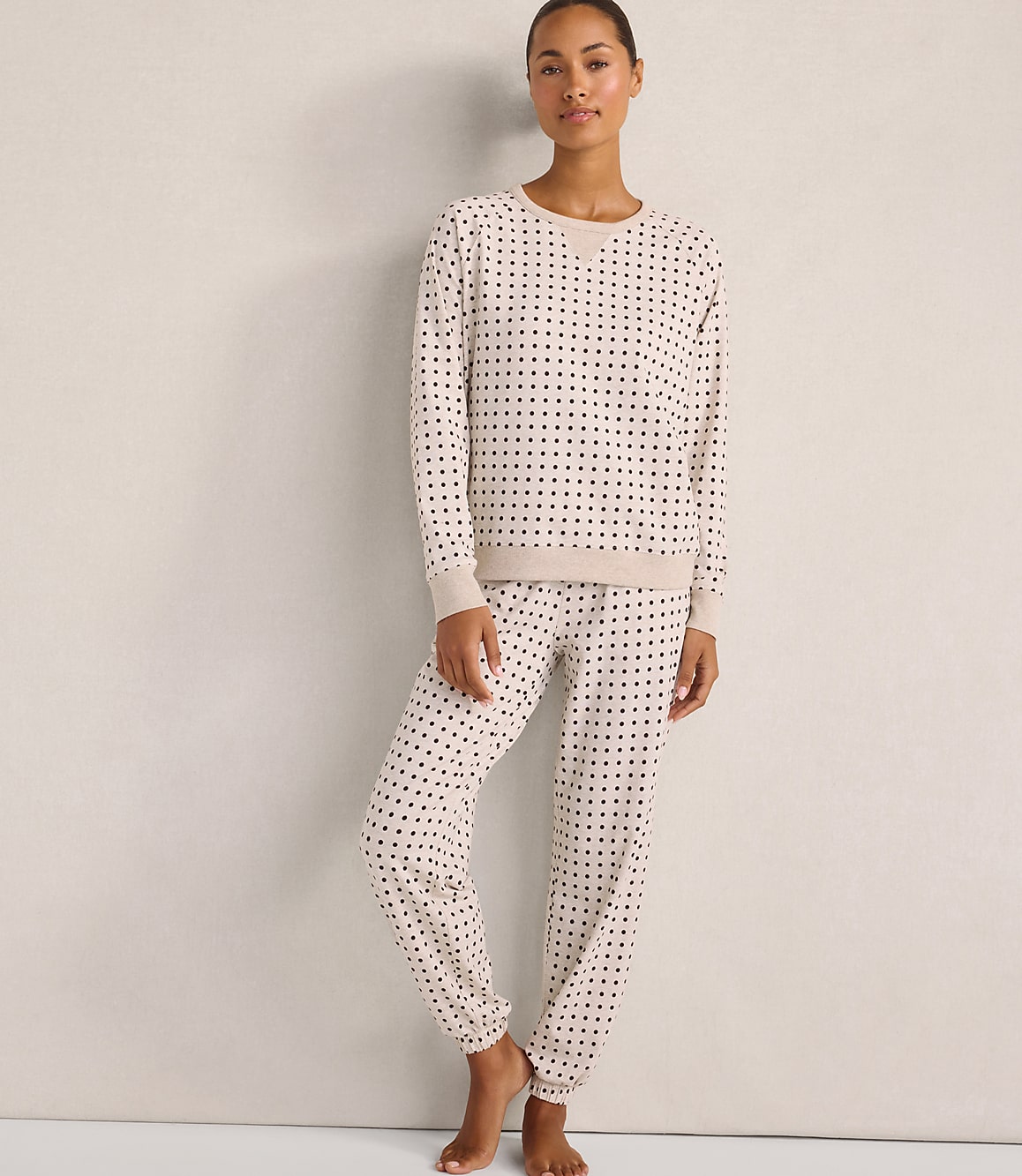 A woman wearing a cream-colored polka-dot pajama set stands against a plain, neutral-toned wall.