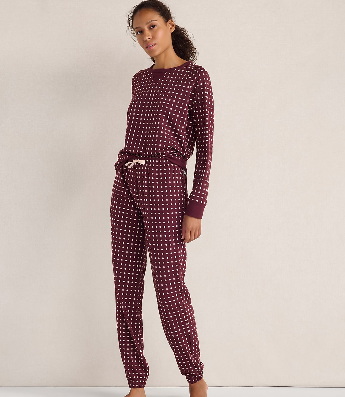 A woman wearing maroon polka-dot pajamas stands against a plain, light-colored wall.