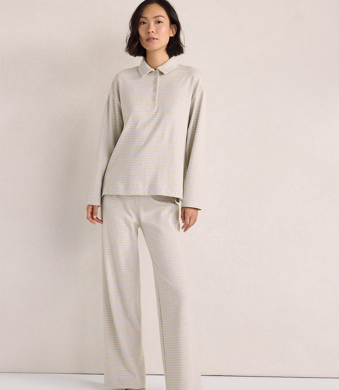 A woman wearing a light-colored, long-sleeved collared shirt and matching wide-leg pants stands against a plain, neutral-toned background.