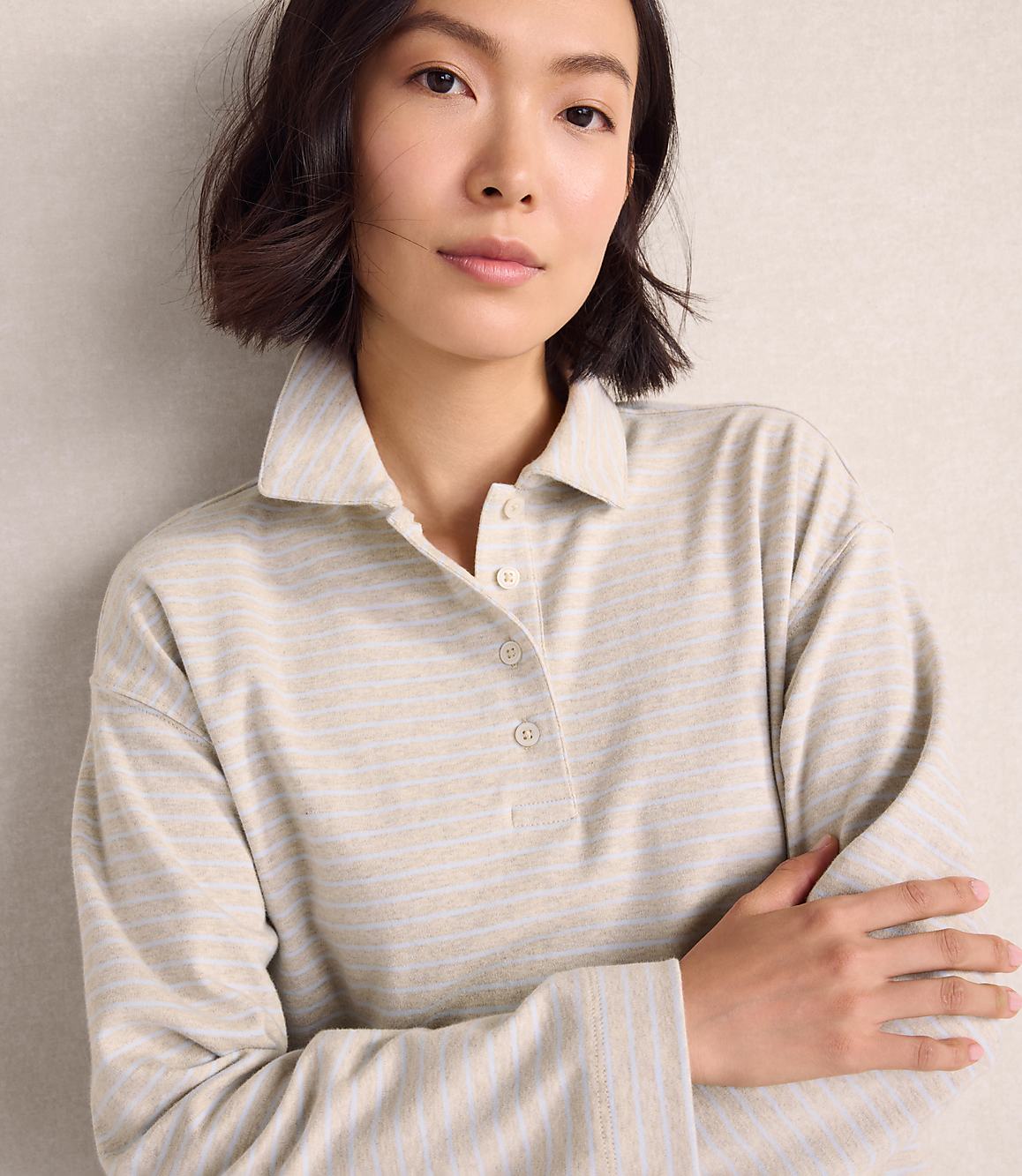 A woman wearing a light-colored striped polo shirt stands with her arms crossed against a plain, neutral-toned wall.