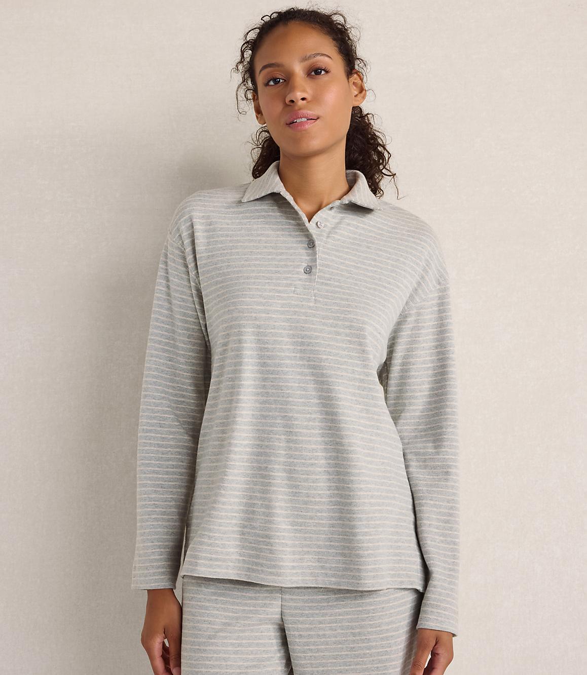 A woman wearing a grey striped long-sleeved polo shirt and matching pants stands against a plain, neutral-colored wall.