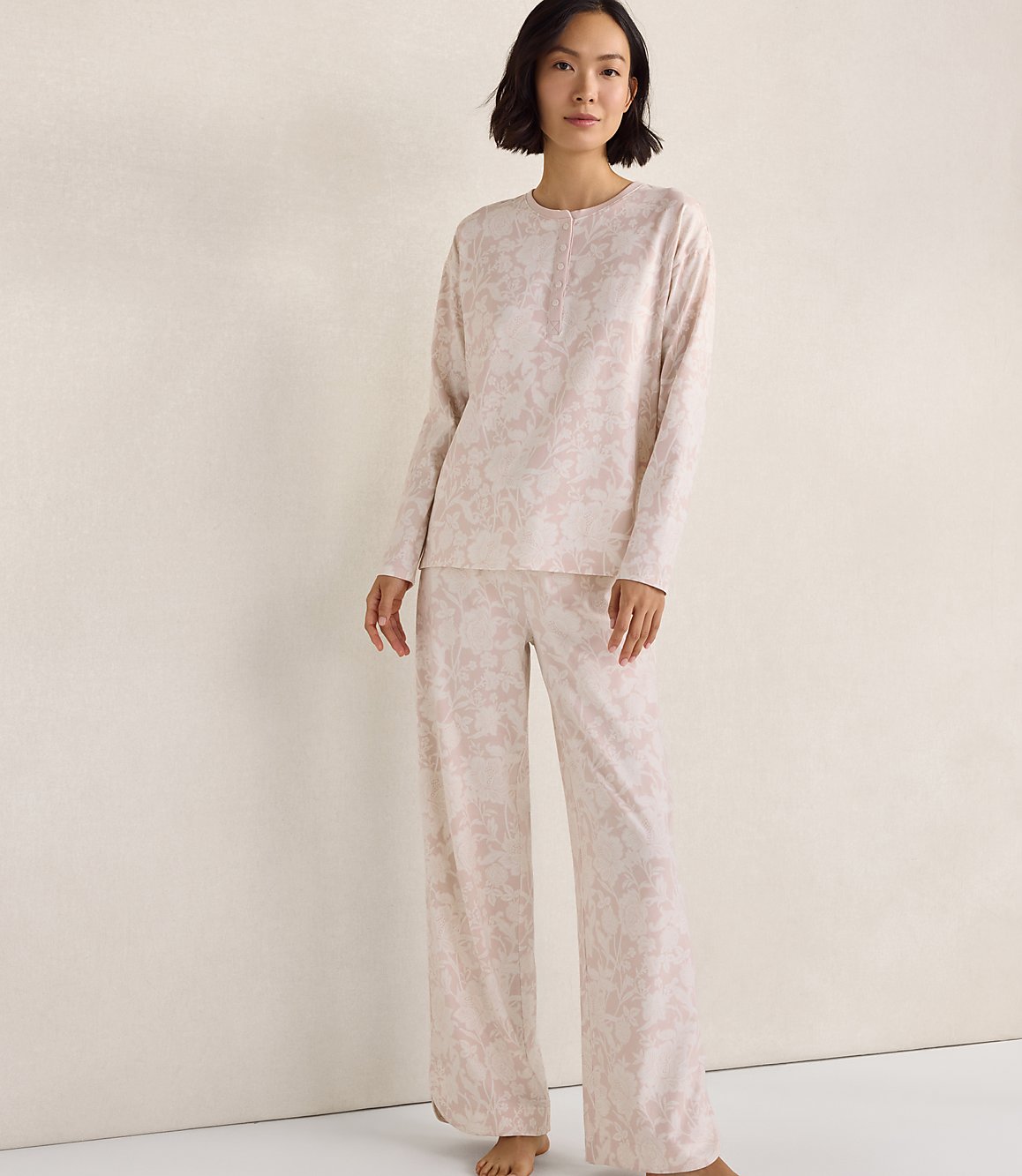 A woman wearing a light pink floral pajama set stands against a plain, neutral-colored wall.