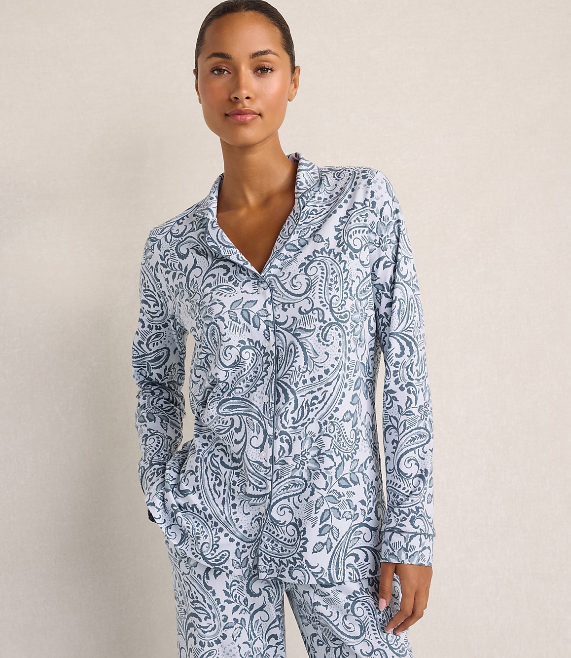 A woman wearing blue and white paisley pajamas stands against a plain, neutral-colored wall.