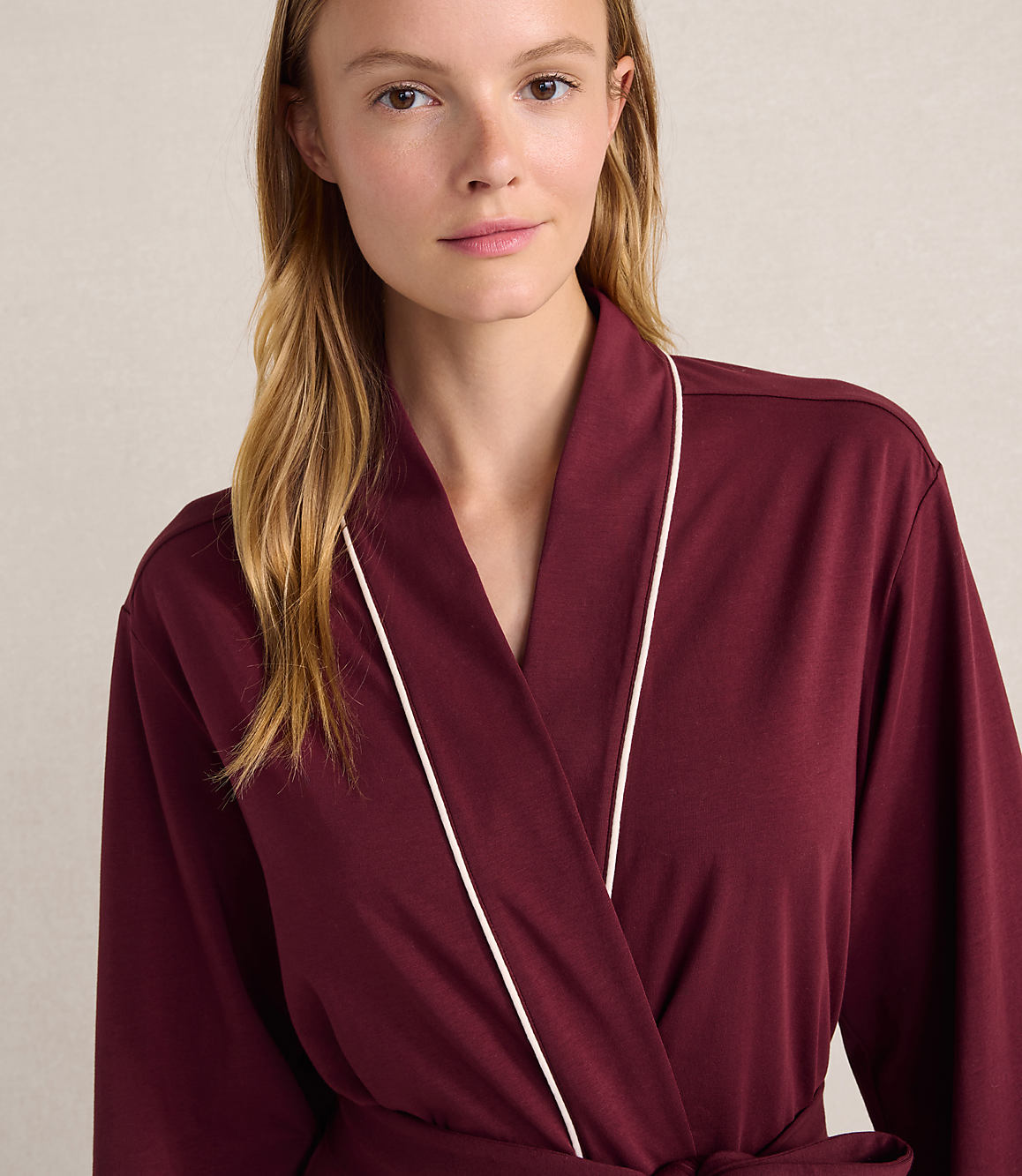 A woman with blonde hair wearing a maroon robe with white trim stands against a plain, light-colored wall.