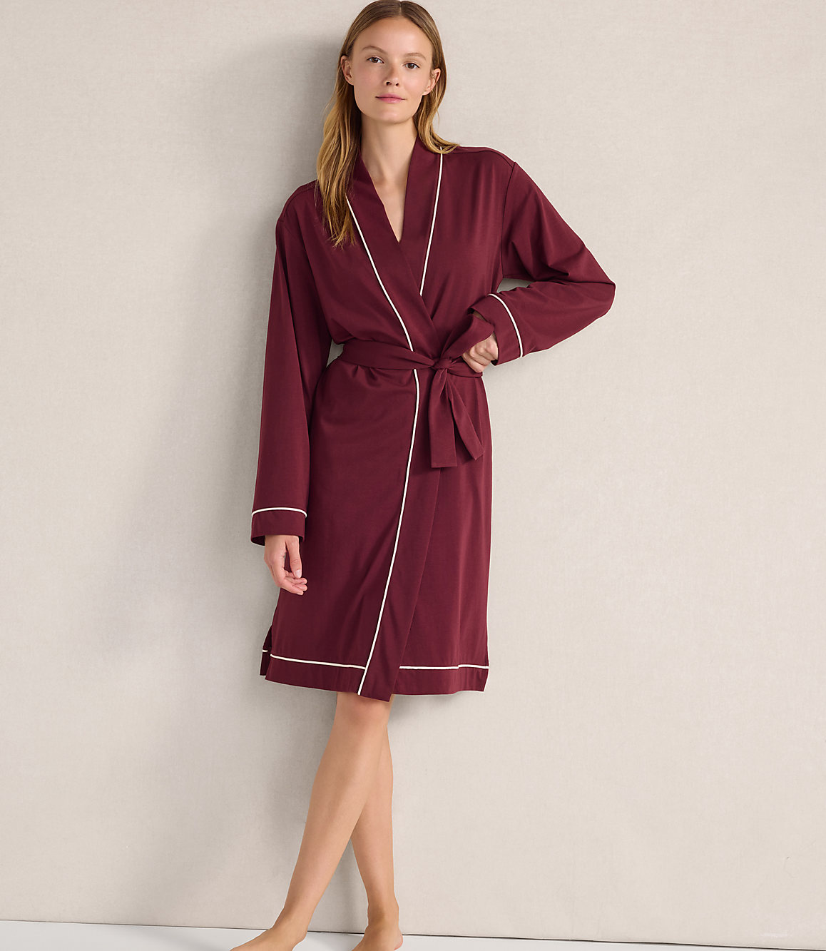 A woman wearing a maroon robe with white trim stands against a plain, light-colored wall.