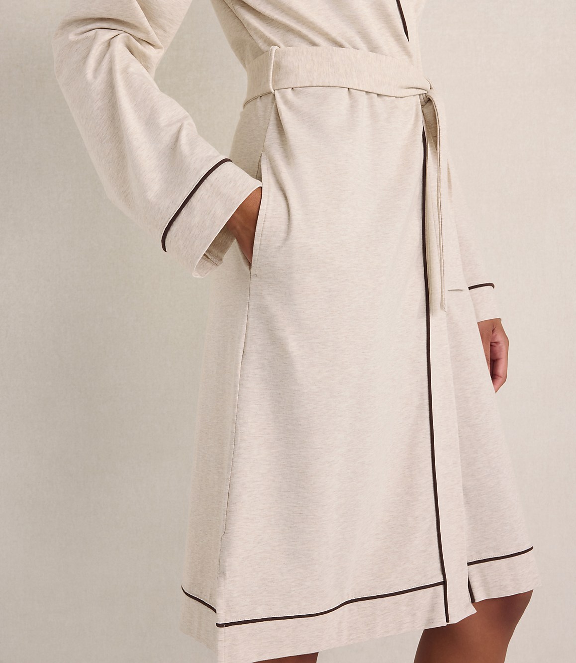 A person wears a cream-colored robe with dark piping against a plain, neutral-toned wall.