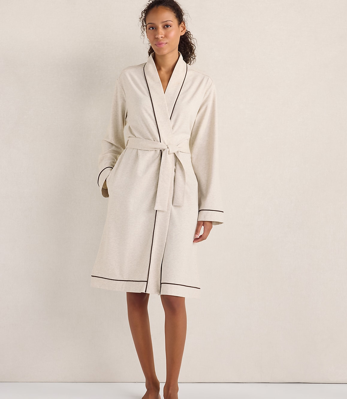 A woman wearing a cream-colored robe with dark piping stands against a plain, neutral-toned wall.