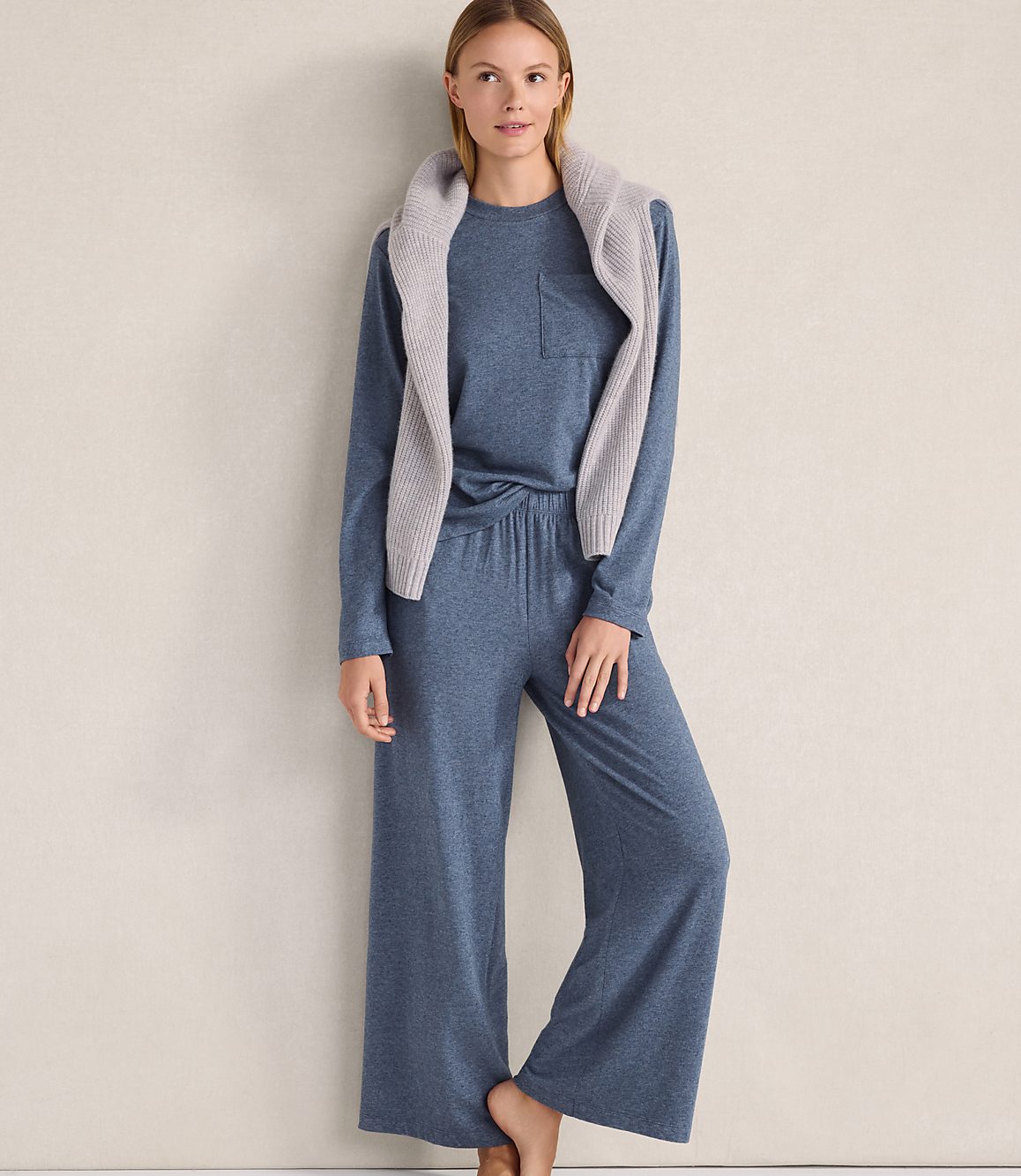 A woman wearing a blue long-sleeved top and wide-leg pants with a grey sweater draped over her shoulders stands against a plain, neutral-colored wall.